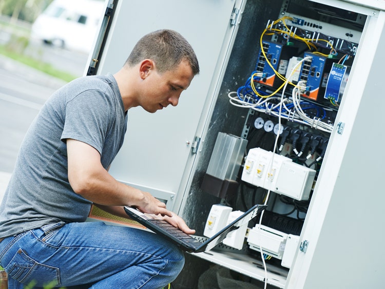 Photo of a male technician outside with a laptop on his lap plugged into a controller in an open HVAC unit